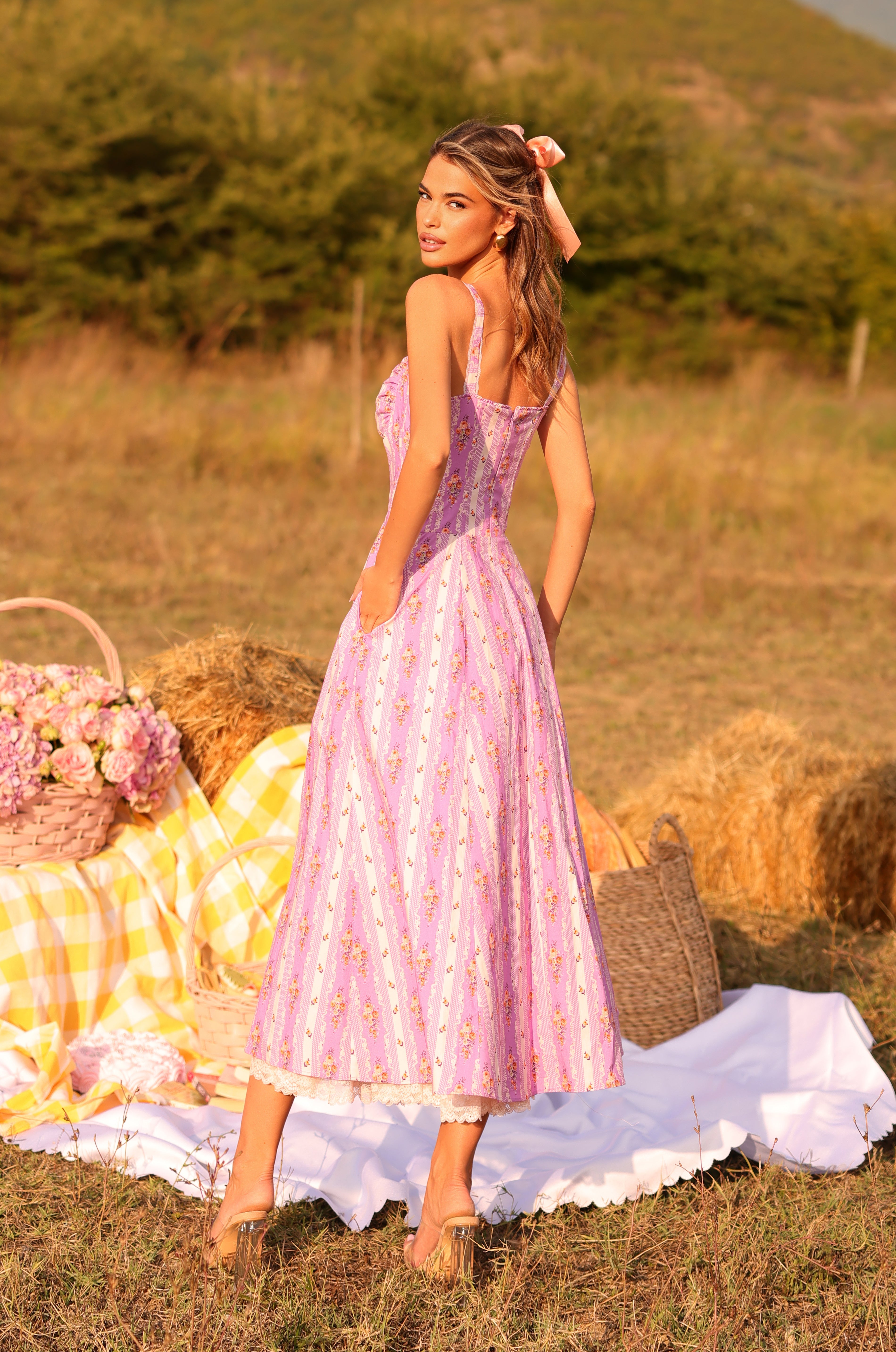 A woman wearing the Lilac Floral Casual Midi Dress: Celenia stands on grass near a picnic setup with flowers, hay bales, and a checkered cloth.
